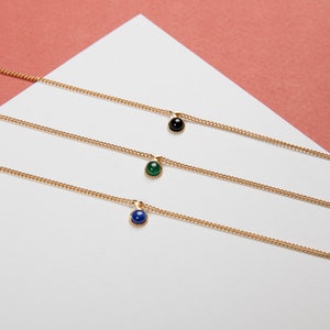 May include: Three delicate gold necklaces, each featuring a small, round gemstone pendant. One pendant is black, one is green, and the third is blue. The necklaces are displayed on a white surface against a pink background.