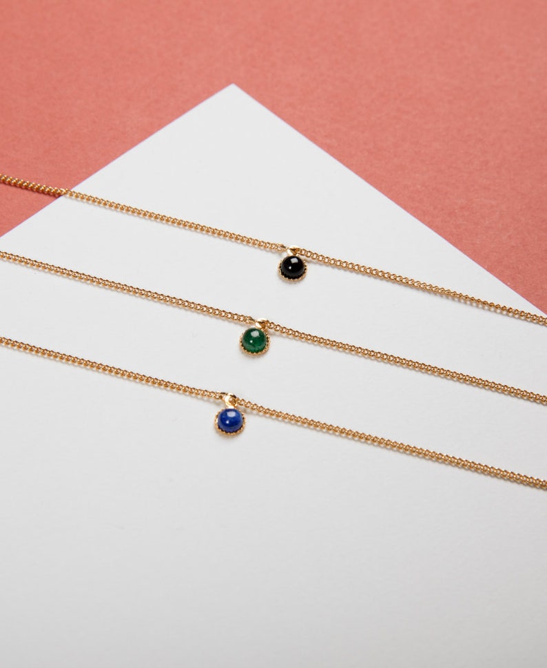May include: Three delicate gold necklaces, each featuring a small, round gemstone pendant. One pendant is black, one is green, and the third is blue. The necklaces are displayed on a white surface against a pink background.