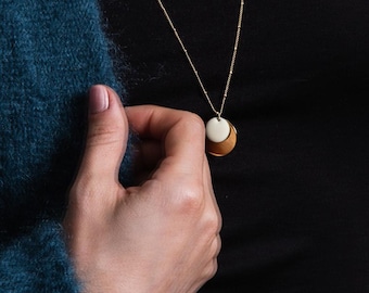 Può includere: Una collana color oro con un ciondolo a forma di disco bianco sovrapposto a una mezzaluna dorata. La collana è su sfondo nero e una mano la tiene. La persona indossa un maglione verde acqua.