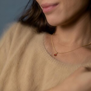 May include: A delicate gold necklace with a small, round, red gemstone pendant. The necklace is worn against a beige sweater. The image is a close-up, focusing on the necklace and the upper chest area.