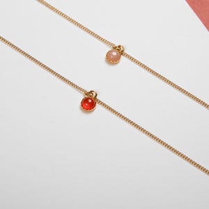 May include: Two delicate gold necklaces, each featuring a small, round gemstone pendant. One pendant is a vibrant red, the other a soft pink. The chains are fine and elegant, perfect for layering or wearing alone. The background is a clean white with a hint of pink.