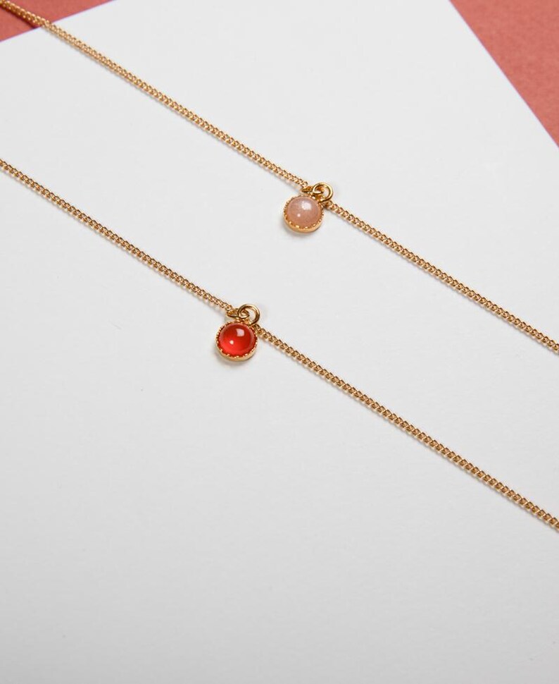 May include: Two delicate gold necklaces, each featuring a small, round gemstone pendant. One pendant is a vibrant red, the other a soft pink. The chains are fine and elegant, perfect for layering or wearing alone. The background is a clean white with a hint of pink.