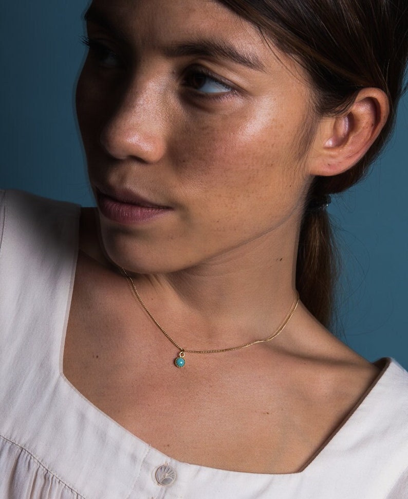 May include: A delicate gold necklace featuring a small, round, turquoise pendant. The necklace is worn against a white top, with a blue background. The pendant is a bright, contrasting color against the gold chain.