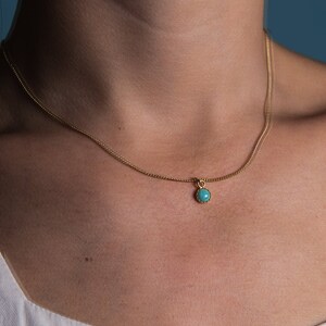 May include: A delicate gold necklace featuring a small, round turquoise pendant. The pendant is set in a gold bezel and hangs from a fine gold chain. The necklace is shown against a neutral background.