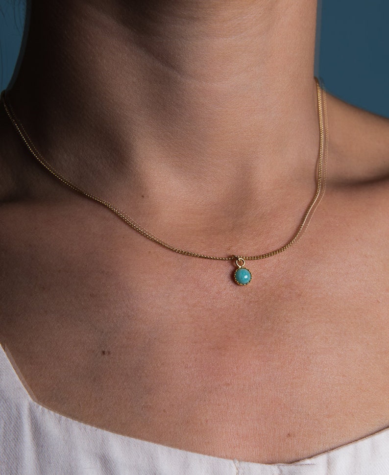 May include: A delicate gold necklace featuring a small, round turquoise pendant. The pendant is set in a gold bezel and hangs from a fine gold chain. The necklace is shown against a neutral background.