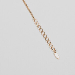 May include: A gold-colored necklace chain with an extender. The chain is composed of small, delicate links, leading to a series of larger, open rings. The extender ends with a small, oval-shaped tag. The background is plain white.