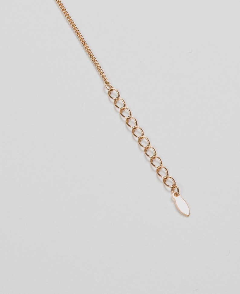May include: A gold-colored necklace chain with an extender. The chain is composed of small, delicate links, leading to a series of larger, open rings. The extender ends with a small, oval-shaped tag. The background is plain white.