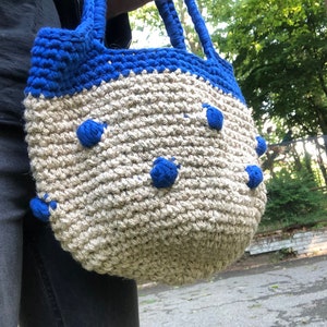 Può includere: Una borsa a mano all'uncinetto con filato blu e beige. La borsa ha dei pompon blu intorno alla parte inferiore.