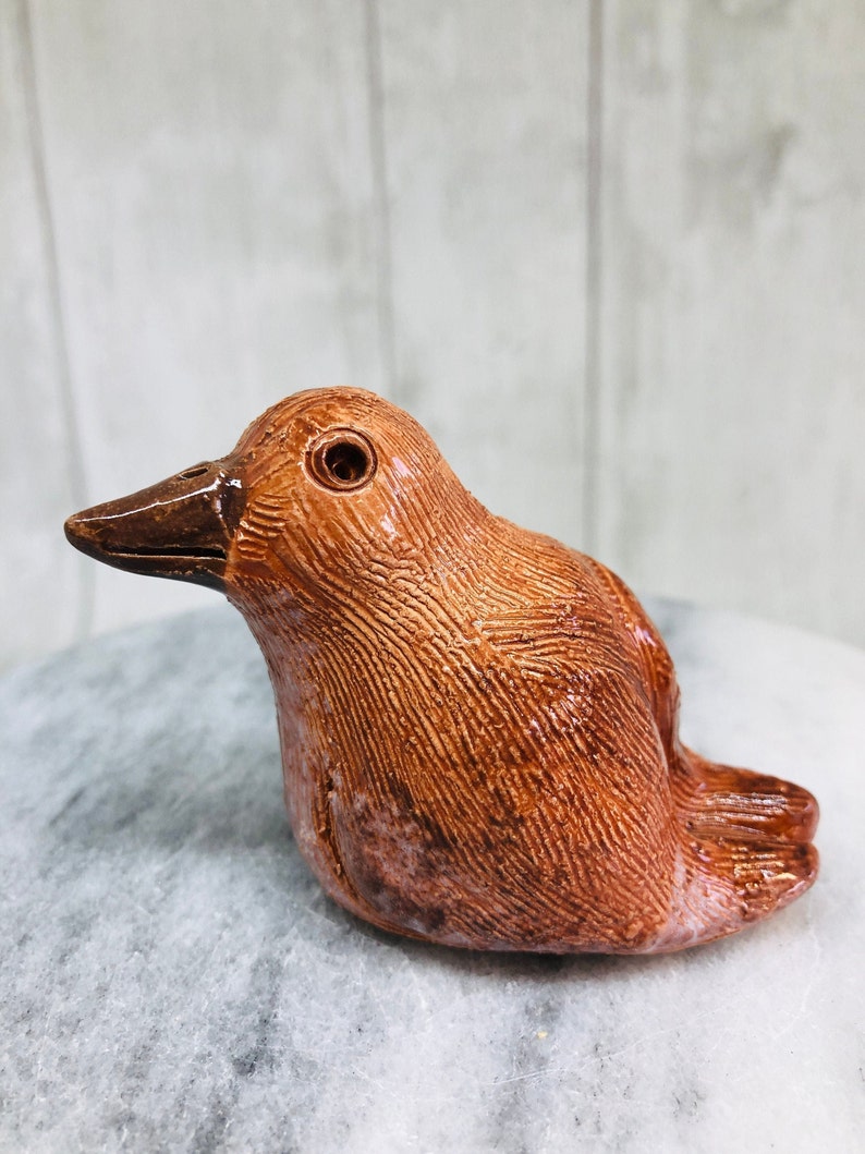 May include: A ceramic bird figurine in shades of brown, with a textured surface. The bird has a dark brown beak and a single visible eye. The figurine is resting on a light gray surface.