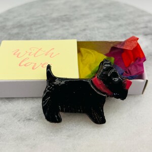 May include: A black Scottie dog figurine with a red collar and nose, presented in an open matchbox. The matchbox has the words "with love" on the front. Colorful tissue paper fills the box.