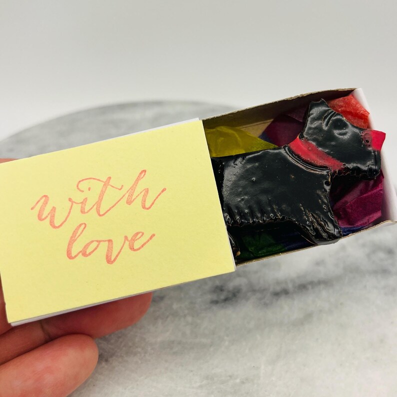 May include: A small matchbox with a yellow front that reads "with love" in cursive. Inside the box is a black dog-shaped item with a red collar, surrounded by colorful tissue paper.