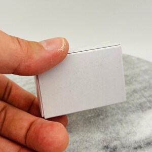 May include: A small, rectangular white box with a closed lid. The box is held in a person's hand, with the fingers partially obscuring the view. The box appears to be made of cardboard or a similar material.