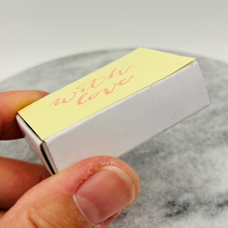 May include: A small, rectangular box with a pale yellow lid and white sides. The lid features the words "with love" in a delicate, cursive script in a soft coral color. The box is held in a hand.