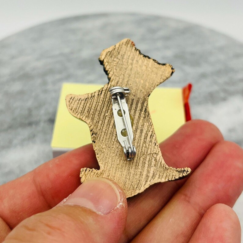 May include: A gold-colored dog-shaped pin with black accents. The pin has a textured surface and a silver pin back. The pin is held in a hand, with a yellow and red object in the background.