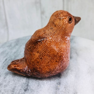 May include: A ceramic bird figurine in shades of brown, with a textured surface and a glossy finish. The bird is perched, with its head turned slightly upwards. The beak is a darker brown, and the eyes are visible.