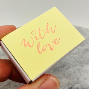 May include: A small, rectangular, yellow box with the words "with love" written in a cursive, coral-colored font. The box is held in a hand.