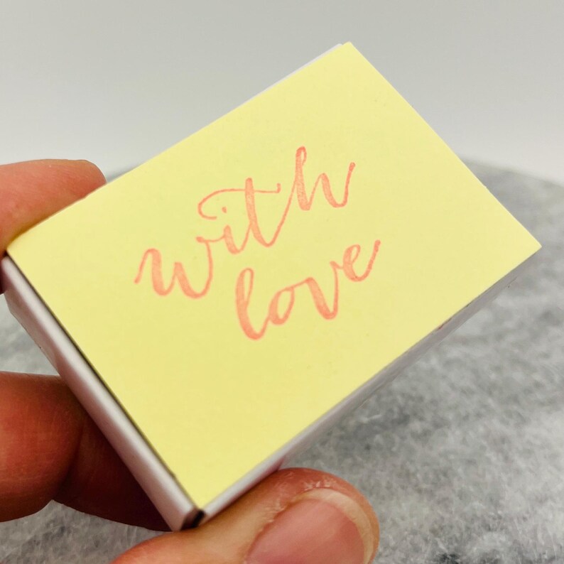 May include: A small, rectangular, yellow box with the words "with love" written in a cursive, coral-colored font. The box is held in a hand.