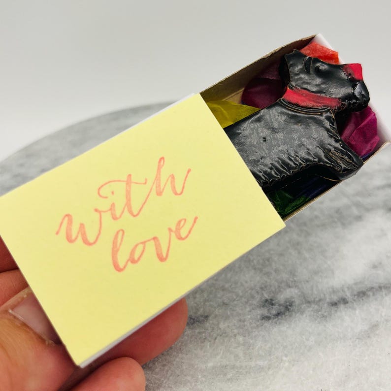 May include: A black dog-shaped wax melt with a red collar rests in a matchbox. The matchbox has a yellow cover with the words "with love" in pink script. The box is lined with colorful tissue paper.