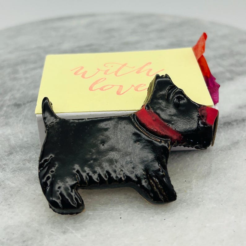 May include: A black Scottie dog figurine with a red collar and nose. The figurine is positioned in front of a yellow card with the words "with love" in pink script. The dog has a glossy finish.