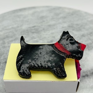 May include: A black Scottie dog figurine with a red collar and nose, resting on a yellow surface. The dog is a stylized representation, likely a decorative item or a collectible. The figurine is set against a neutral background.