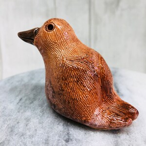 May include: A ceramic bird figurine in shades of brown, with a textured surface and a glossy finish. The bird has a dark brown beak and eye, and is perched on a gray surface. The figurine is a decorative item.