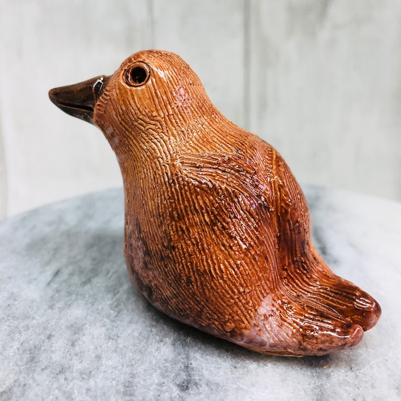 May include: A ceramic bird figurine in shades of brown, with a textured surface and a glossy finish. The bird has a dark brown beak and eye, and is perched on a gray surface. The figurine is a decorative item.