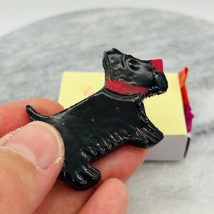 May include: A black Scottie dog figurine with a red collar and red accents on the head. The dog is glossy and appears to be made of a hard material. The figurine is held in a hand, with a yellow and white box in the background.
