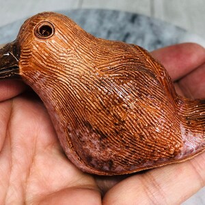 May include: A ceramic bird figurine with a textured, brown surface and a dark brown beak. The bird has a small, round eye and is held in a hand. The figurine is likely a decorative item or collectible.