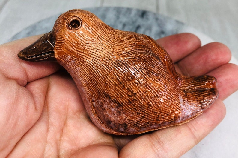 May include: A ceramic bird figurine with a textured, brown surface and a dark brown beak. The bird has a small, round eye and is held in a hand. The figurine is likely a decorative item or collectible.