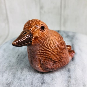 May include: A handcrafted ceramic bird figurine in shades of brown. The bird has a detailed textured surface, a dark brown beak, and a single eye. This decorative piece is a charming addition to any home decor collection.