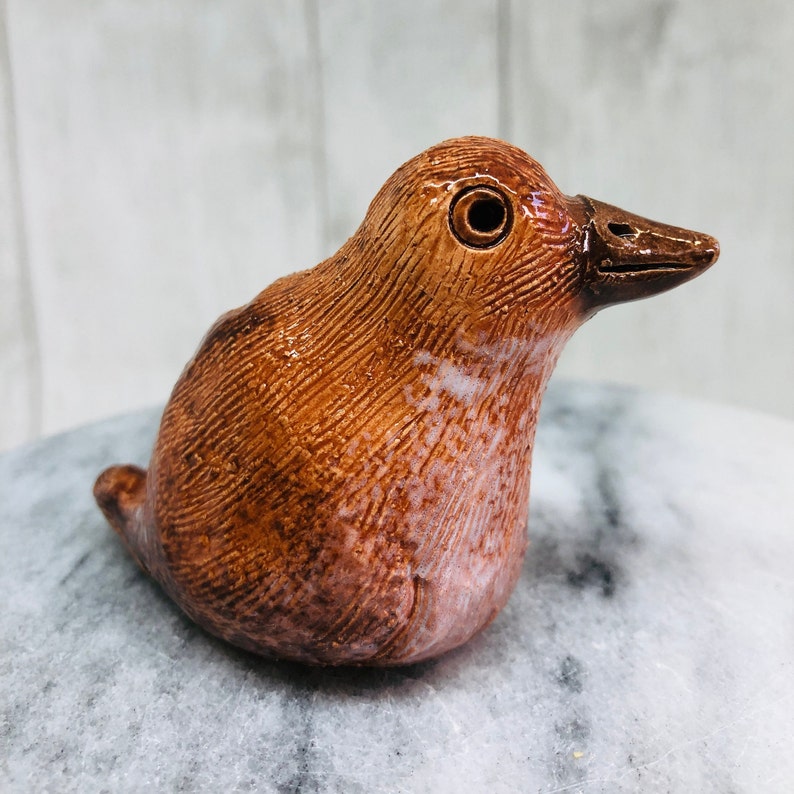 May include: A ceramic bird figurine in shades of brown, with a textured surface. The bird has a dark brown beak and a round eye. The figurine is sitting on a gray surface.