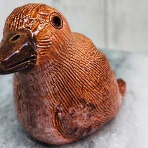May include: A ceramic duck figurine in shades of brown. The duck has a detailed textured surface with a glossy finish on the beak and eye. The figurine is a decorative item.