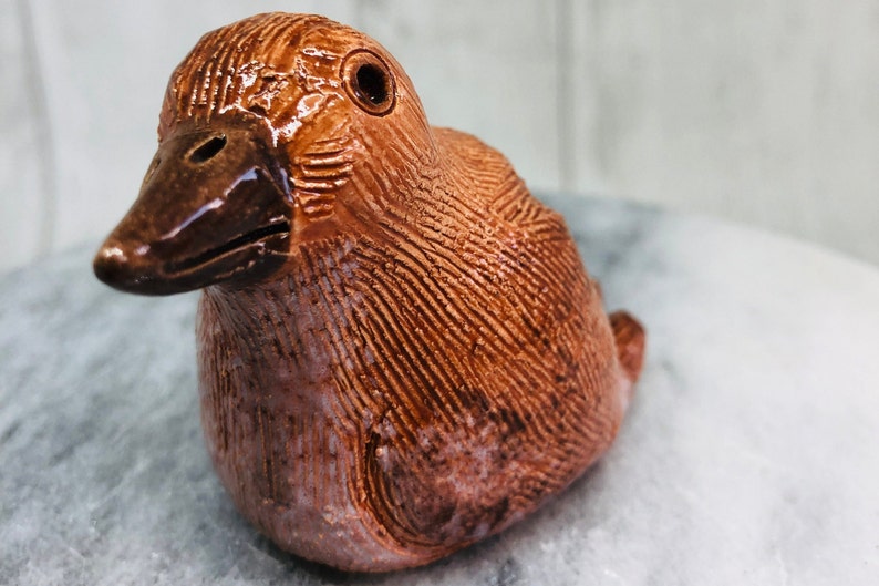 May include: A ceramic duck figurine in shades of brown. The duck has a detailed textured surface with a glossy finish on the beak and eye. The figurine is a decorative item.
