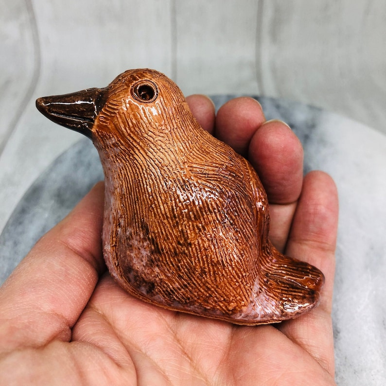 May include: A ceramic bird figurine with a textured, brown surface and a glossy finish. The bird has a dark brown beak and eye detail. The figurine is held in a hand.