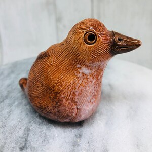 May include: A handcrafted ceramic bird figurine in shades of brown and tan. The bird has a textured surface with detailed lines, a dark brown beak, and a single eye. The figurine is a unique decorative item.