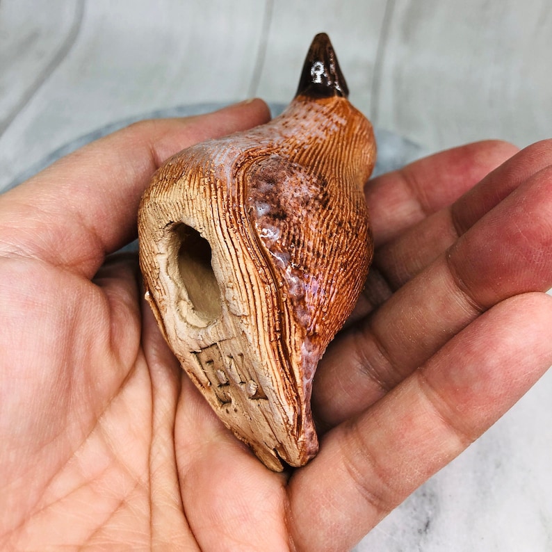 May include: A handmade ceramic bird figurine in shades of brown, tan, and white. The bird has a textured surface with a small opening and a dark brown beak. The figurine is held in a person's hand.