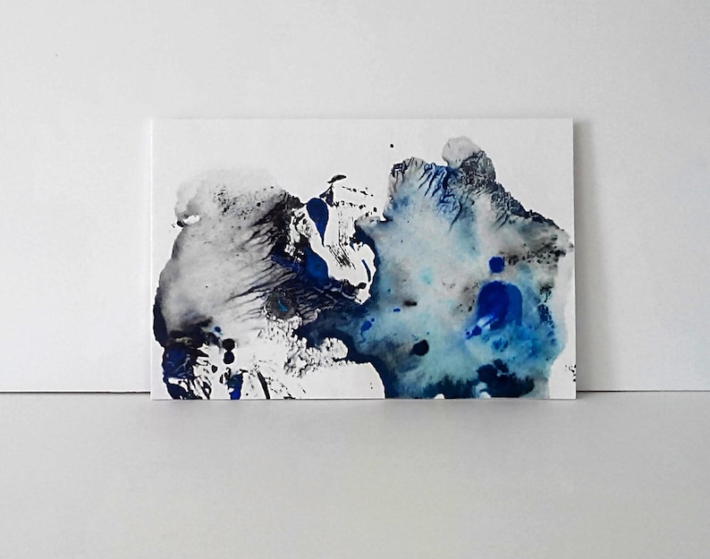 May include: Abstract art piece featuring a blend of blue, black, and gray ink on a white canvas. The artwork showcases a dynamic interplay of colors and textures, with fluid shapes and splatters. The piece is mounted on a white surface.