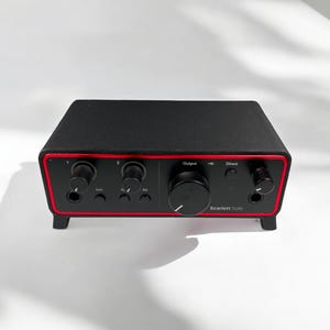 May include: A black audio interface with a red trim border, featuring multiple circular control knobs, input ports, and buttons on the front panel. The text Scarlett Solo is printed on the lower right side, with labels for Output, Direct, and other settings.