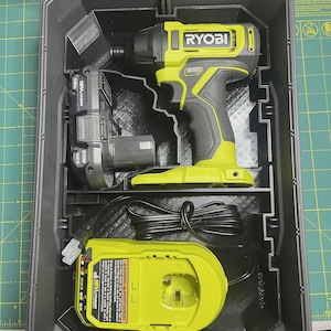 May include: A lime green and gray Ryobi impact driver, an 18V lithium battery, and a battery charger with a black power cord, all organized inside a dark gray plastic carrying case with molded compartments for each tool component.
