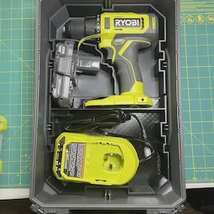 May include: A lime green and gray Ryobi cordless drill with a battery pack and a battery charger stored inside a gray plastic carrying case. The drill features a black chuck, a trigger, and a battery slot. The charger has a black power cord.