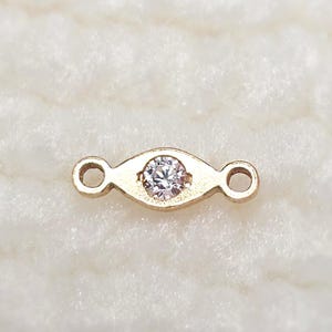 May include: A gold-colored jewelry connector with a clear, round gemstone in the center. The connector has an oval shape with a loop on each end. The gemstone is set in a bezel setting.