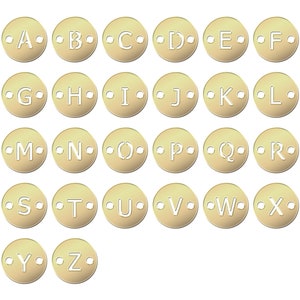 May include: A collection of gold-colored metal alphabet charms, each with a letter from A to Z. The letters are cut out, and each charm has two small holes for attachment. The charms are arranged on a white background.
