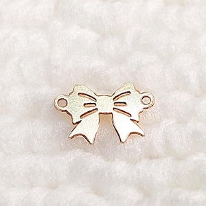 May include: A gold-coloured bow charm with a cut-out design. The charm has two small holes on either side, likely for attaching to a necklace or bracelet. The bow is centered on a white, textured background.