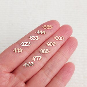 Może przedstawiać: Kolekcja małych, złotych zawieszek z numerami, w tym 111, 222, 333, 444, 555, 000, 777, 888 i 999. Te zawieszki są idealne do tworzenia biżuterii lub projekt&oacute;w rzemieślniczych, na jasnym tle.