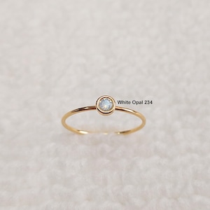 May include: A delicate gold ring featuring a small, round white opal gemstone. The ring has a thin band and a simple, elegant design. The text "White Opal 234" is visible.