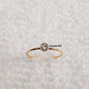 May include: A delicate gold ring featuring a single, clear crystal gemstone set in a bezel. The ring has a slender band and the text "Crystal 001" is visible.