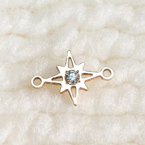 May include: A gold-colored charm in the shape of a compass rose with a central, iridescent gemstone. The charm has two small loops on either side for attachment. The background is a soft, white, textured surface.