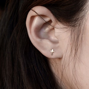 May include: A close-up of an ear with a small, gold cross stud earring. The earring has a simple, minimalist design. The person's skin is fair, and their dark hair is visible in the background.