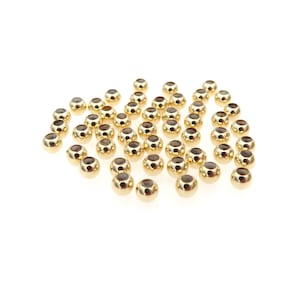 May include: A collection of small, gold-colored beads with a dark center, scattered on a white surface. The beads have a shiny, metallic finish and a rounded shape, suitable for jewelry making or craft projects.