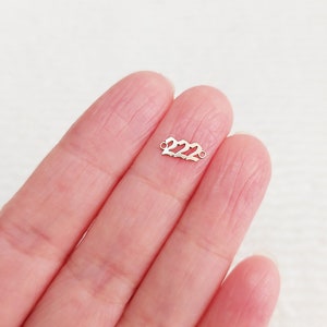 Może przedstawiać: Mały, srebrny charms z wyciętą liczbą "222". Charms ma dwie małe pętle na każdym końcu do mocowania do biżuterii. Charms jest trzymany w dłoni osoby.